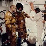 Astronaut Lousma is assisted with a glove adjustment to flight suit